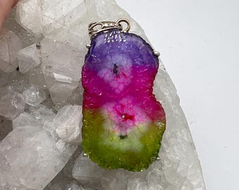 Peut inclure: Un pendentif composé d'une tranche de pierre d'agate avec un dégradé de couleurs violet, rose vif et vert citron. La pierre présente une forme naturelle irrégulière avec une texture cristalline et est fixée par une bélière en métal argenté.
