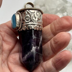 May include: An amethyst crystal pendant with a silver-coloured metal cap and bail. The cap has an ornate design with embossed details and a small blue stone. The crystal is a deep purple colour with white veining.