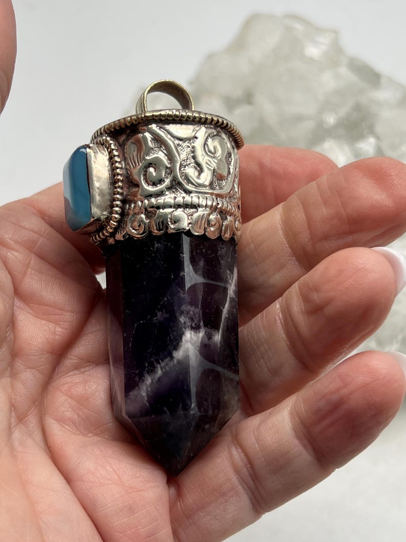 May include: An amethyst crystal pendant with a silver-coloured metal cap and bail. The cap has an ornate design with embossed details and a small blue stone. The crystal is a deep purple colour with white veining.