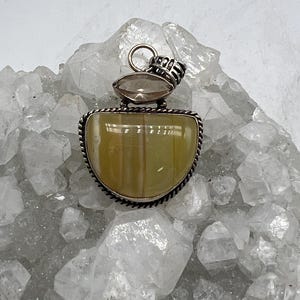 May include: A silver pendant with a large, light yellow-green, semi-circular stone. The stone is set in a silver frame with a twisted rope design. Above the main stone is a smaller, oval-shaped, brown stone. The pendant has a silver bail.