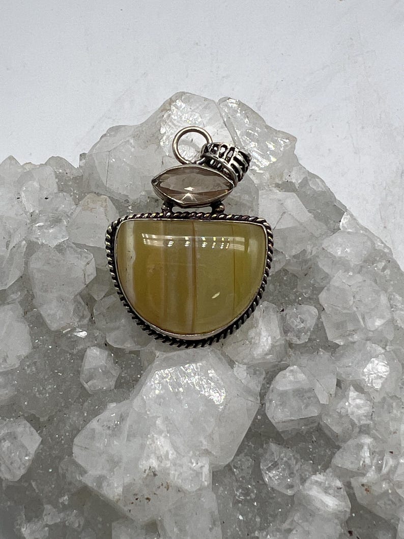 May include: A silver pendant with a large, light yellow-green, semi-circular stone. The stone is set in a silver frame with a twisted rope design. Above the main stone is a smaller, oval-shaped, brown stone. The pendant has a silver bail.