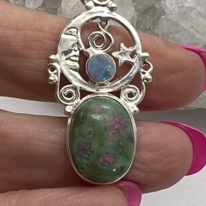 May include: A silver pendant with a crescent moon design, a star, and a blue opal stone. The pendant is attached to a green and pink gemstone.