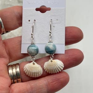 May include: Dangle earrings featuring round, marbled blue and white beads paired with small, off-white ribbed seashells. The components are connected by silver-toned metal findings and suspended from silver hooks on a white display card labeled Sterling Silver.