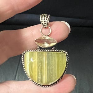 May include: A silver pendant with a large, light green, semi-circular stone, accented by a smaller, oval, yellow stone. The pendant has a decorative silver bail and a rope-like border around the main stone. The pendant is held by a hand.