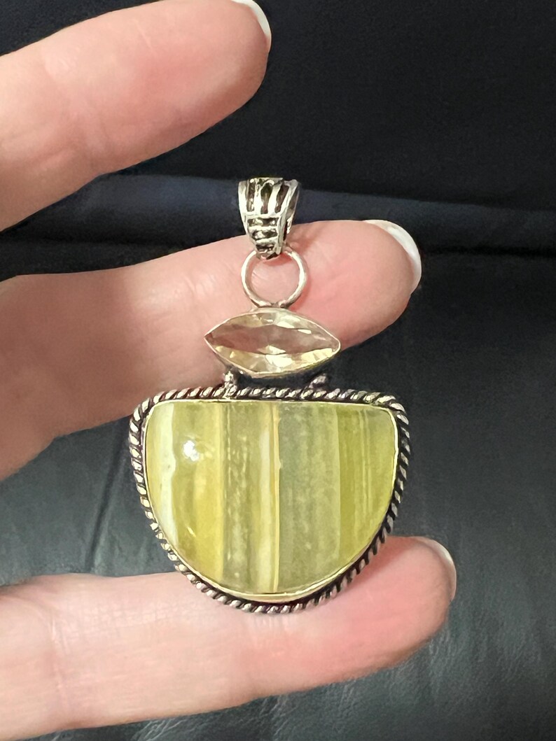 May include: A silver pendant with a large, light green, semi-circular stone, accented by a smaller, oval, yellow stone. The pendant has a decorative silver bail and a rope-like border around the main stone. The pendant is held by a hand.