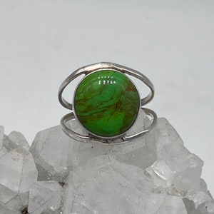 May include: A silver ring featuring a large, round, vibrant green stone with brown veining. The ring has a simple, elegant design with a double band on each side of the stone. The ring is displayed on a white, crystalline surface.