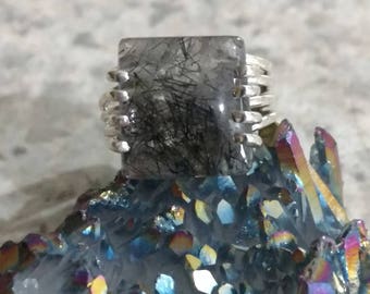 Può includere: Un anello d'argento con una grande pietra rettangolare trasparente contenente inclusioni nere. L'anello è incastonato su un gruppo di cristalli iridescenti con sfumature blu, viola e gialle. La fascia dell'anello ha un design strutturato.