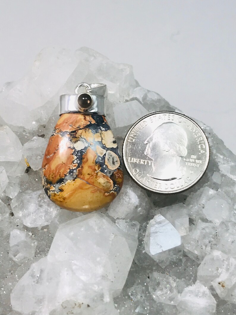 May include: A pendant with a silver bail and a teardrop-shaped stone. The stone features a mix of tan, brown, and blue patterns. A quarter is next to the pendant for size comparison.