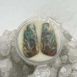 Peut inclure: Deux pierres précieuses polies avec des motifs turquoise, rouges et beiges sont présentées dans un récipient en plastique transparent. Les pierres ont une forme rectangulaire aux bords arrondis. Le fond est un amas de roches cristallines blanches.