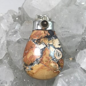 May include: A teardrop-shaped pendant with a silver bail and a small, dark brown stone. The pendant features a mosaic pattern of tan, brown, black, and white colors. The stone is set in a silver bezel.