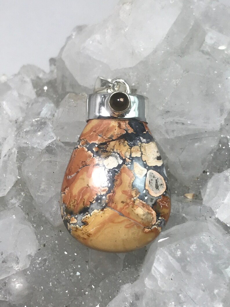 May include: A teardrop-shaped pendant with a silver bail and a small, dark brown stone. The pendant features a mosaic pattern of tan, brown, black, and white colors. The stone is set in a silver bezel.