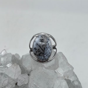 May include: A silver ring with a white and black dendritic agate stone. The ring is set on a bed of white crystals.