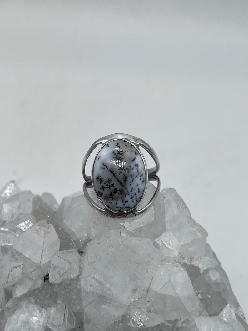 May include: A silver ring with a white and black dendritic agate stone. The ring is set on a bed of white crystals.