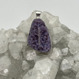 May include: A pendant featuring a unique, lavender-colored druzy stone with a silver-tone bail. The stone has a bubbly texture and a natural, organic shape. The pendant is displayed on a bed of clear quartz crystals.