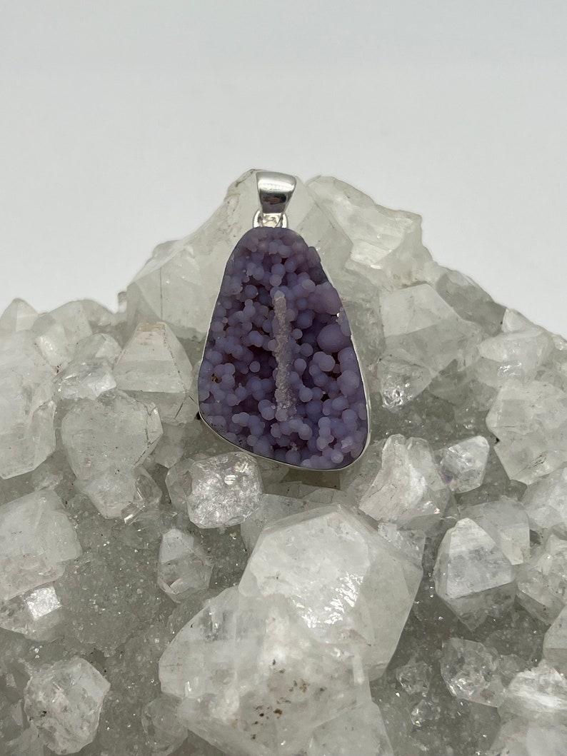 May include: A pendant featuring a unique, lavender-colored druzy stone with a silver-tone bail. The stone has a bubbly texture and a natural, organic shape. The pendant is displayed on a bed of clear quartz crystals.