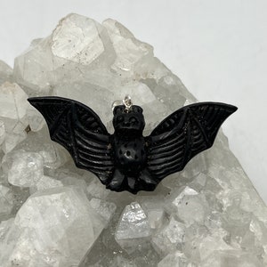 May include: Black carved bat pendant with a silver bail. The pendant is sitting on a bed of white crystals.