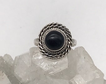 May include: A silver ring featuring a large, circular, black gemstone. The stone is encircled by a twisted rope pattern. The ring is set against a white backdrop.