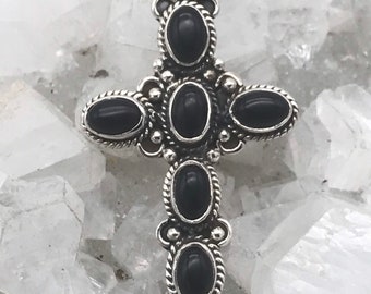 Puede incluir: Un colgante de cruz de plata con cinco piedras ovaladas negras. La cruz está detallada con pequeñas cuentas de plata alrededor de los bordes de las piedras preciosas.