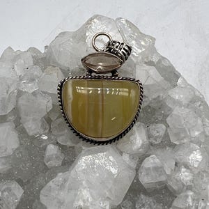 May include: A silver pendant with a light yellow, half-moon shaped stone, framed by a twisted silver border. A smaller, oval-shaped, light brown stone sits above the larger stone. The pendant has a silver loop for a chain.