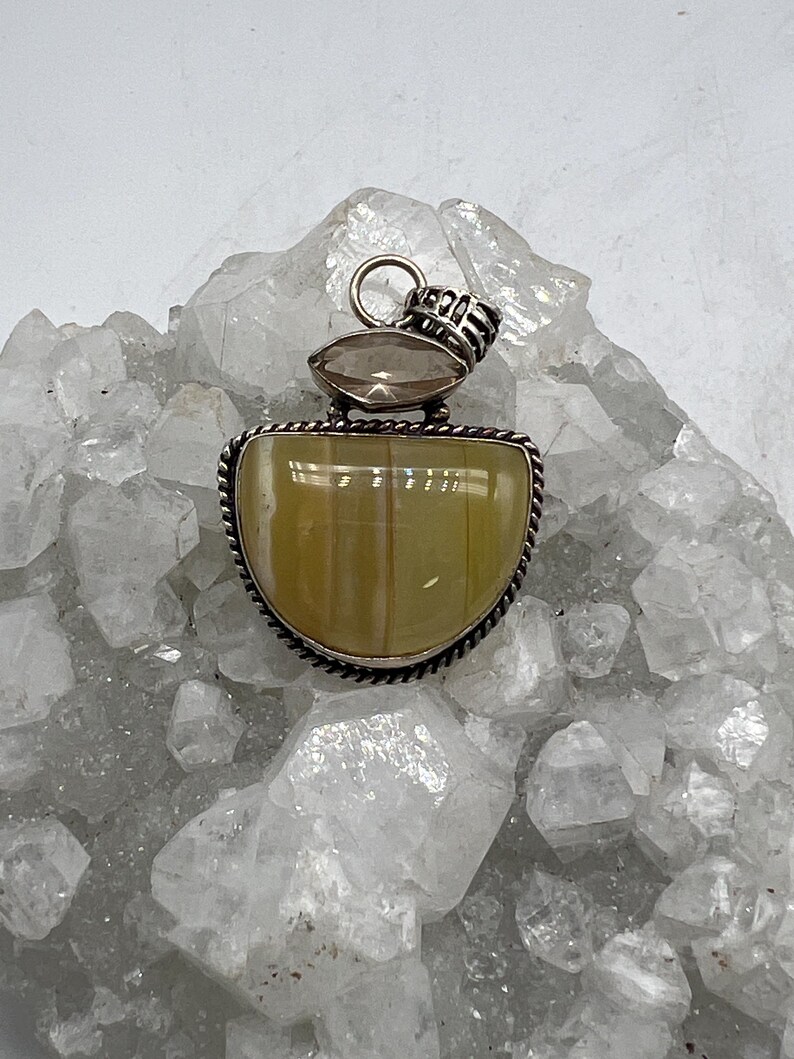 May include: A silver pendant with a light yellow, half-moon shaped stone, framed by a twisted silver border. A smaller, oval-shaped, light brown stone sits above the larger stone. The pendant has a silver loop for a chain.