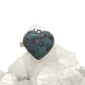 May include: A silver ring featuring a heart-shaped green and pink gemstone. The gemstone has a speckled pattern.