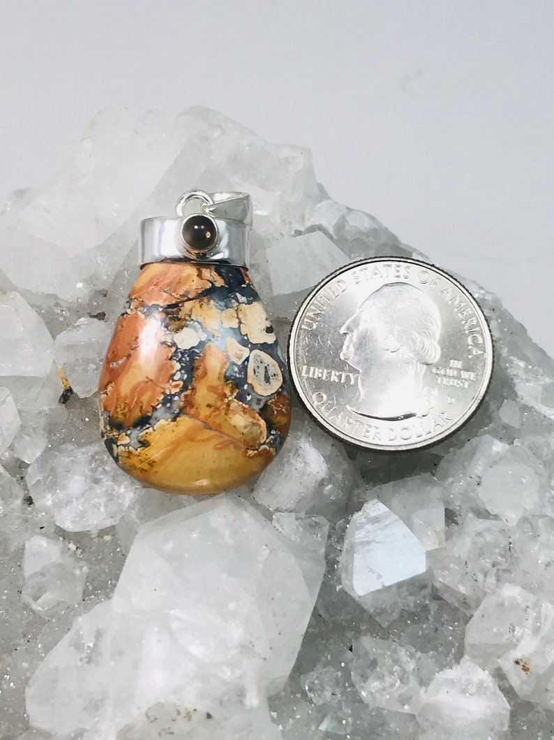 May include: A teardrop-shaped pendant with a silver bail and a small brown stone. The pendant features a mix of tan, brown, and white patterns. A U.S. quarter is next to the pendant for size comparison. The pendant is likely a piece of jewelry.