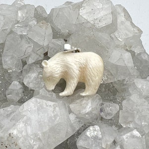 May include: A cream-colored bear pendant with a silver-toned metal bail attached to the top. The bear is carved with visible fur texture and is positioned in a walking stance, facing toward the left side of the frame.