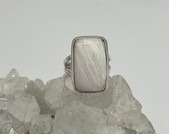 May include: A rectangular, white gemstone ring with a silver band. The gemstone has a layered, textured appearance. The ring is set on a white, crystalline surface, with a plain white background.