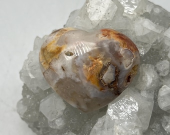 Puede incluir: Una piedra en forma de corazón con tonos beige, naranja y blanco, posada sobre un grupo de cristales transparentes. Este corazón de piedra natural es un objeto decorativo. Los cristales son claros y facetados.
