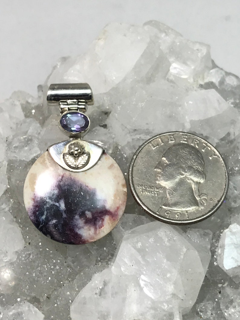 May include: A round, white and purple stone pendant with a silver bail and a small amethyst gemstone. The pendant is about the size of a quarter.