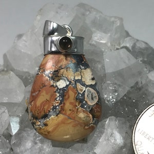 May include: A teardrop-shaped pendant with a silver bail. The pendant features a mosaic pattern of tan, brown, gray, and white colors. A small, dark brown stone is set in the silver bail. The pendant is displayed on a bed of clear crystals.