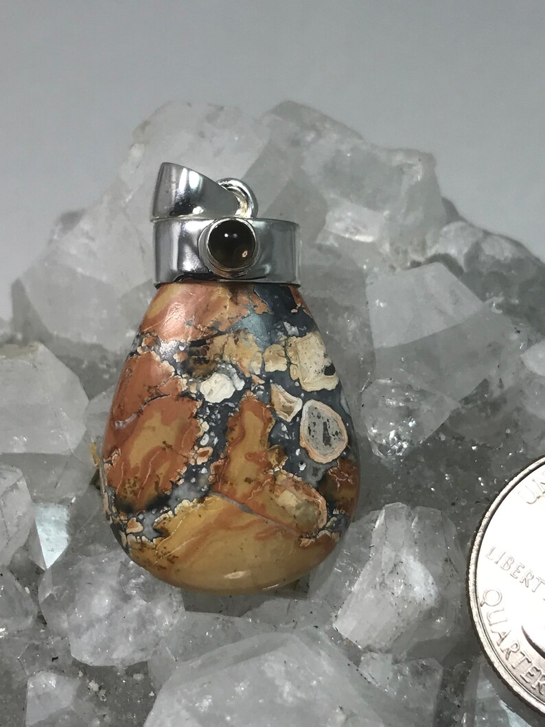 May include: A teardrop-shaped pendant with a silver bail. The pendant features a mosaic pattern of tan, brown, gray, and white colors. A small, dark brown stone is set in the silver bail. The pendant is displayed on a bed of clear crystals.