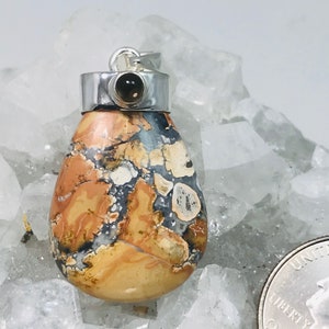May include: A teardrop-shaped pendant with a silver bail and a small, dark gemstone. The pendant features a patterned stone with shades of tan, brown, gray, and white. The pendant is displayed on a white, crystalline surface.