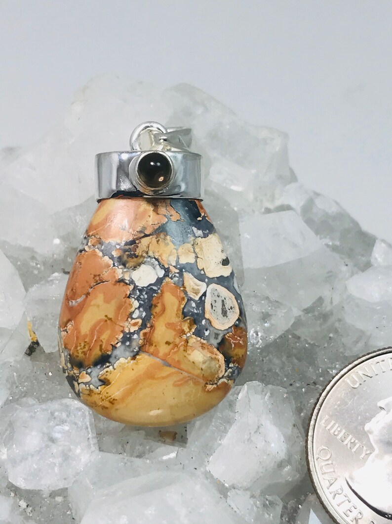May include: A teardrop-shaped pendant with a silver bail and a small, dark gemstone. The pendant features a patterned stone with shades of tan, brown, gray, and white. The pendant is displayed on a white, crystalline surface.