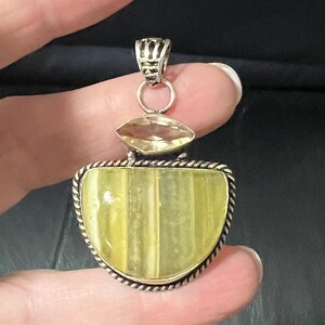 May include: A silver pendant with a large, light yellow, half-moon shaped stone. The stone is set in a silver frame with a twisted rope design. Above the main stone is a smaller, oval-shaped yellow stone. The pendant is held by a silver bail.