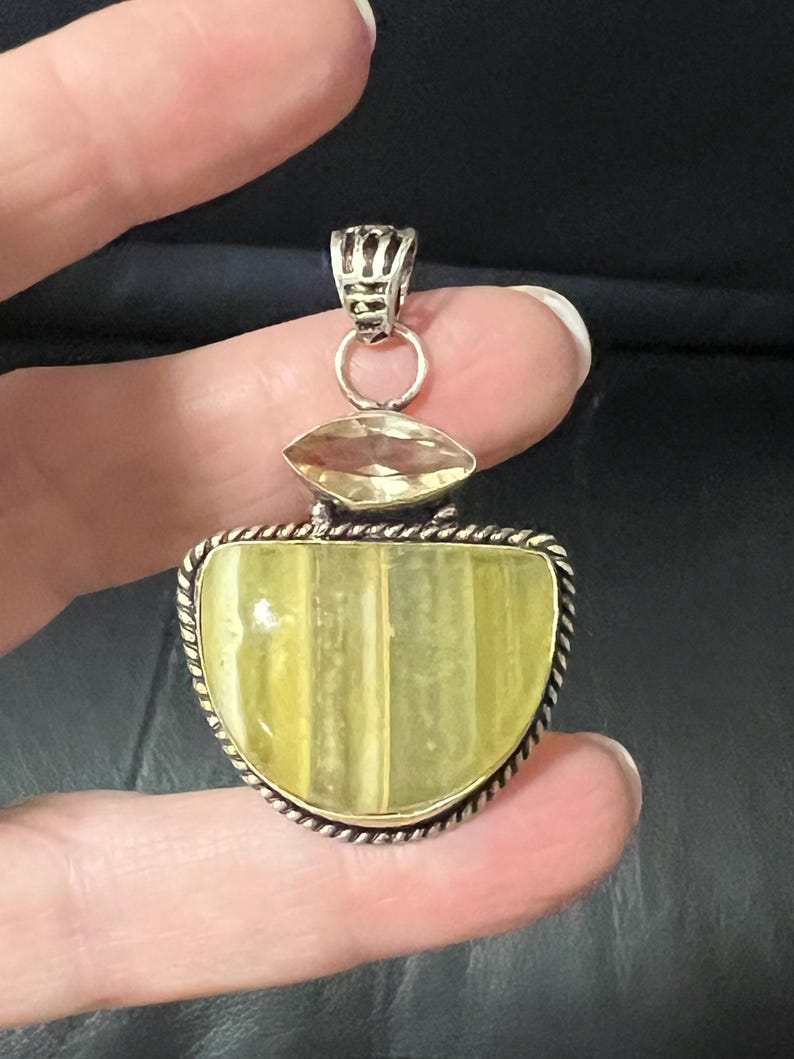 May include: A silver pendant with a large, light yellow, half-moon shaped stone. The stone is set in a silver frame with a twisted rope design. Above the main stone is a smaller, oval-shaped yellow stone. The pendant is held by a silver bail.