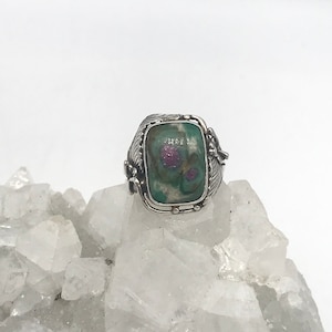 Könnte beinhalten: Ein silberner Ring mit einem grünen und rosafarbenen Edelstein, der in eine detaillierte Silberfassung gefasst ist. Der Ring liegt auf einem Bett aus weißen Kristallen.