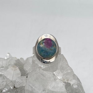 Peut inclure: Une bague en argent ornée d'une grosse pierre ovale. La pierre présente un mélange de couleurs, notamment du bleu sarcelle, du vert et du rouge vif. La bague a un anneau en argent simple et poli, et la pierre est sertie dans un sertissage à lunette.
