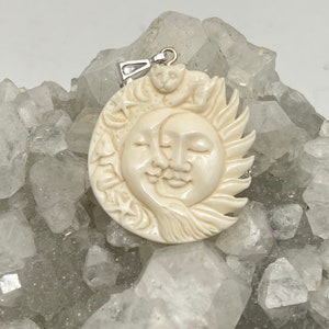 May include: A white carved pendant featuring a sun and moon face with a small cat on top. The pendant is made of bone and has a silver bail.