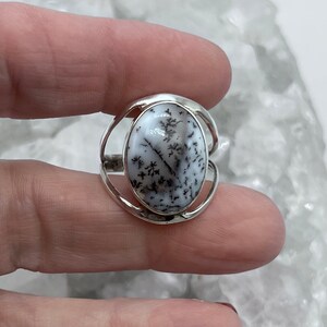 May include: A silver ring with an oval-shaped dendritic agate stone. The stone is white with black dendritic patterns.