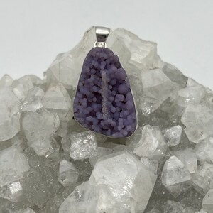 May include: A pendant featuring a polished, grape-like, purple chalcedony stone set in a silver-colored metal. The stone has a bubbly texture and a natural, irregular shape. The pendant is displayed on a bed of clear quartz crystals.