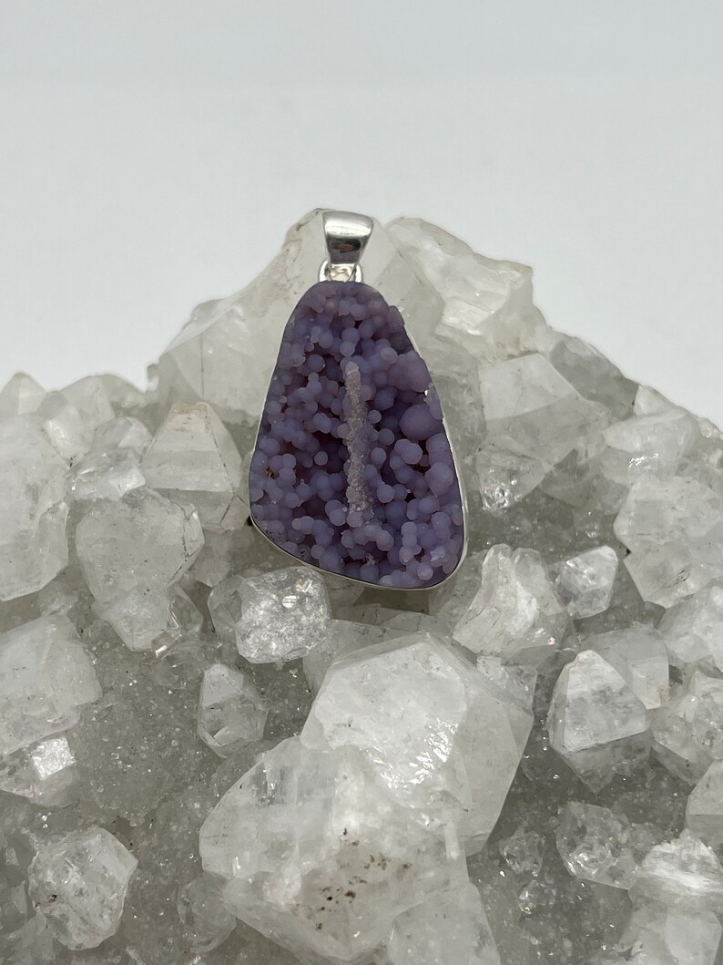 May include: A pendant featuring a polished, grape-like, purple chalcedony stone set in a silver-colored metal. The stone has a bubbly texture and a natural, irregular shape. The pendant is displayed on a bed of clear quartz crystals.