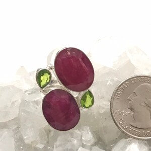 May include: A silver ring featuring two large oval-cut, deep red gemstones and two smaller pear-shaped, light green gemstones. A U.S. quarter coin is included for size comparison. The ring is set against a white, crystalline background.