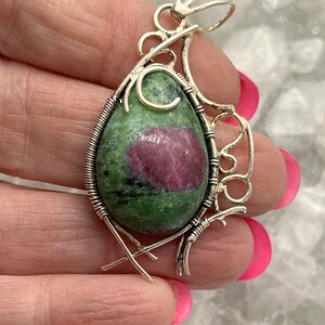 May include: A handcrafted pendant featuring a green and red gemstone, wire-wrapped in a silver-colored metal. The gemstone has a teardrop shape. The pendant has an ornate design with decorative swirls and cross-like accents.