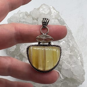 May include: A silver pendant with a large, half-moon shaped, yellow-toned stone. The pendant has a smaller, oval-shaped stone above the larger stone. The pendant is held by a hand against a white, crystalline background.