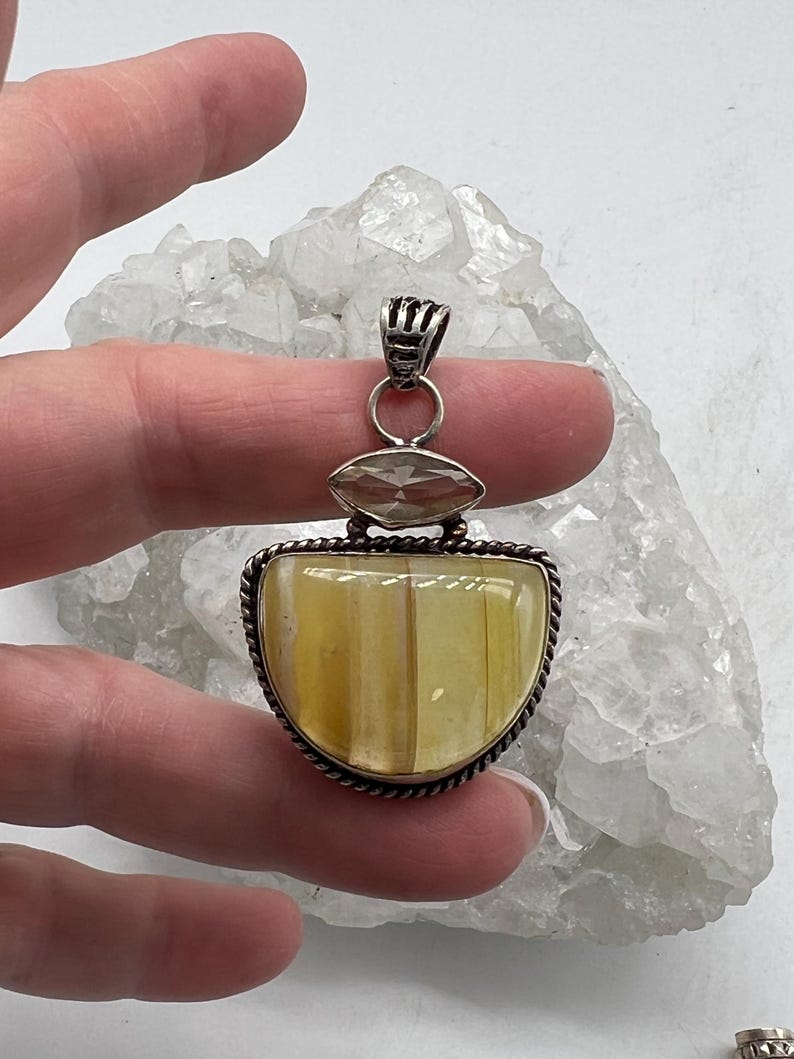 May include: A silver pendant with a large, half-moon shaped, yellow-toned stone. The pendant has a smaller, oval-shaped stone above the larger stone. The pendant is held by a hand against a white, crystalline background.