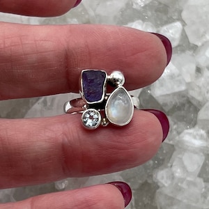 Può includere: Un anello in argento sterling con un gruppo di pietre preziose. L'anello presenta un'ametista viola rettangolare, una pietra di luna a forma di pera, un topazio blu rotondo e una piccola sfera d'argento. Le pietre sono incastonate.