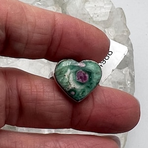 May include: A heart-shaped ring featuring a green and white stone with a central red inclusion. The ring has a silver band. The stone's pattern resembles a natural landscape. The ring is held up against a white background.