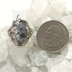 Pu&ograve; includere: Un anello d'argento con una pietra preziosa bianca e nera incastonata in una montatura d'argento. L'anello &egrave; decorato con spirali e foglie d'argento. L'anello &egrave; mostrato accanto a una moneta da 25 centesimi di dollaro americano per il confronto delle dimensioni.