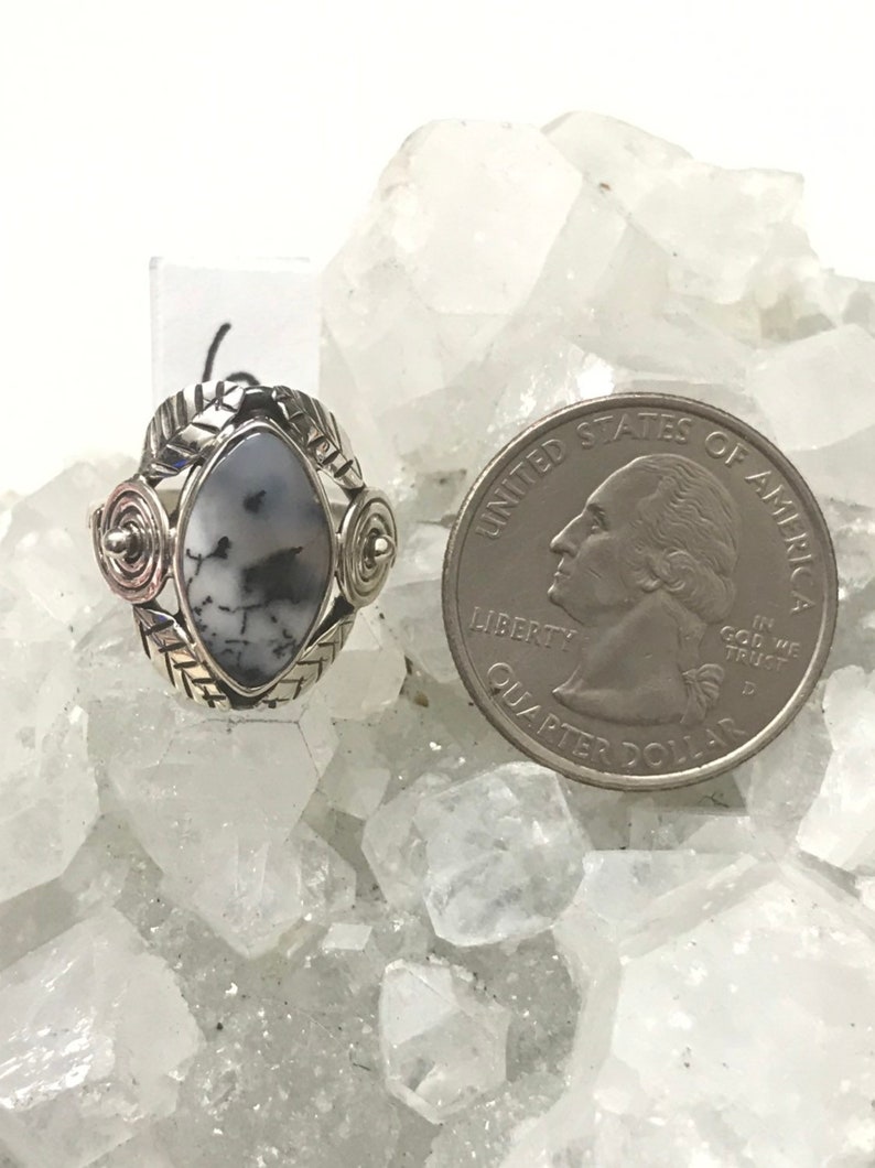 Pu&ograve; includere: Un anello d'argento con una pietra preziosa bianca e nera incastonata in una montatura d'argento. L'anello &egrave; decorato con spirali e foglie d'argento. L'anello &egrave; mostrato accanto a una moneta da 25 centesimi di dollaro americano per il confronto delle dimensioni.